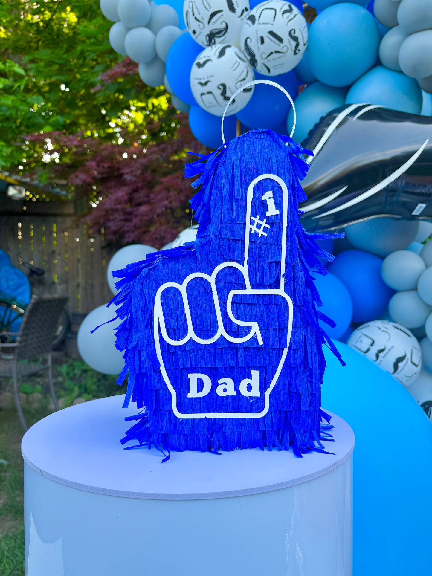 Blue foam finger piñata with 'Dad' on a white pedestal against a backdrop of balloons.