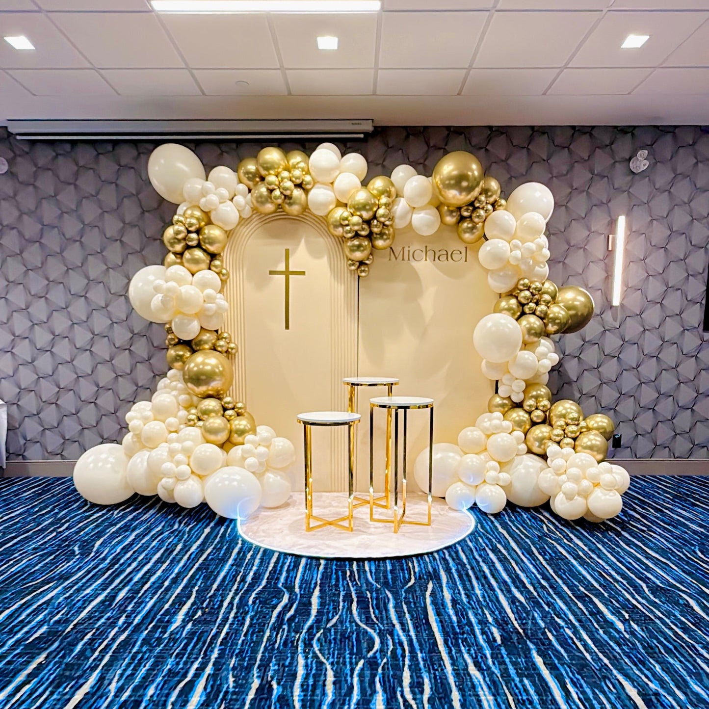 Decorative setup with balloons, a cross, and candles on a blue carpeted floor.