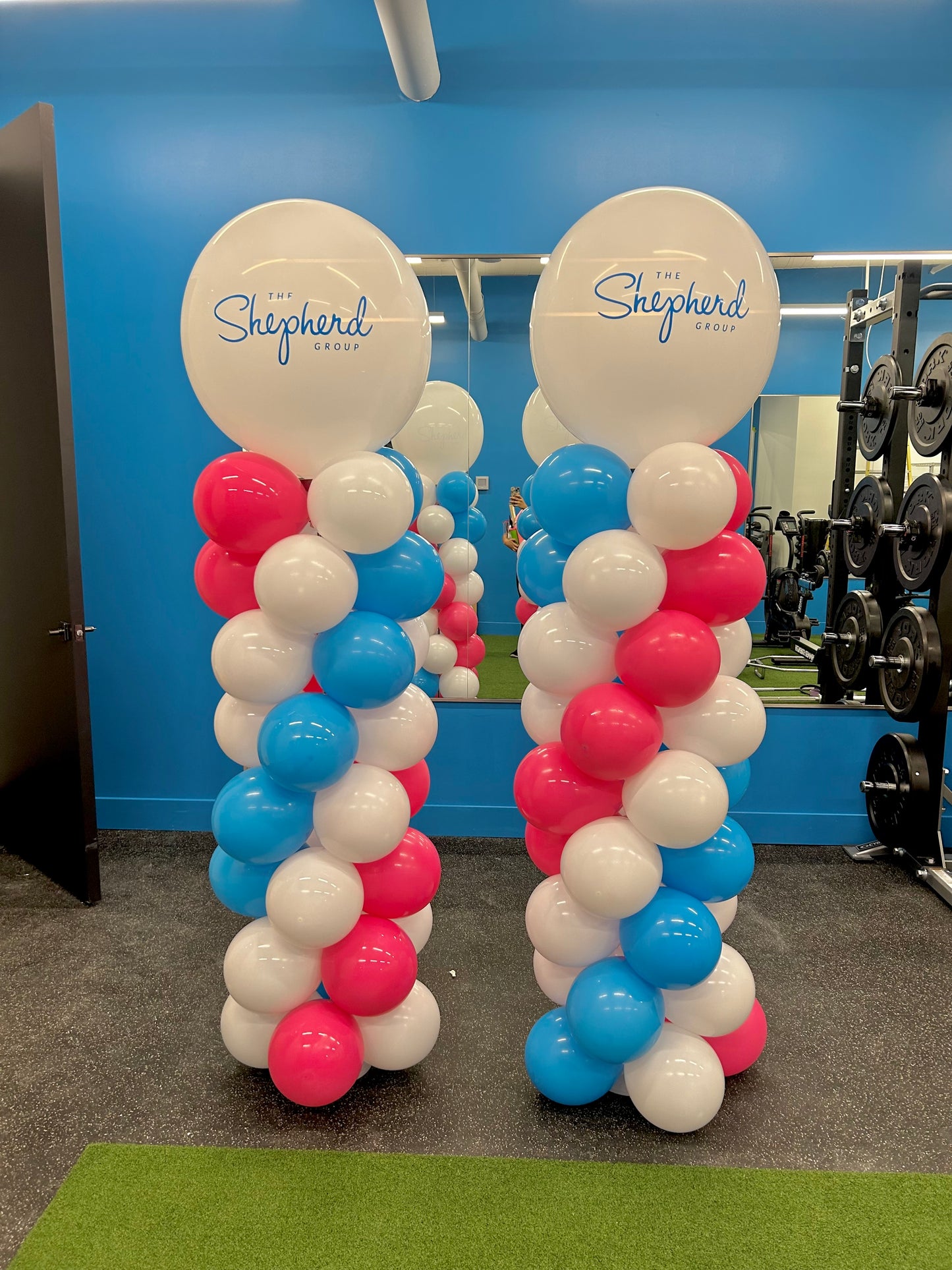 Two freestanding balloon columns at an indoor event, featuring a pattern of red, white, and blue balloons with a logo printed on the top white balloon.