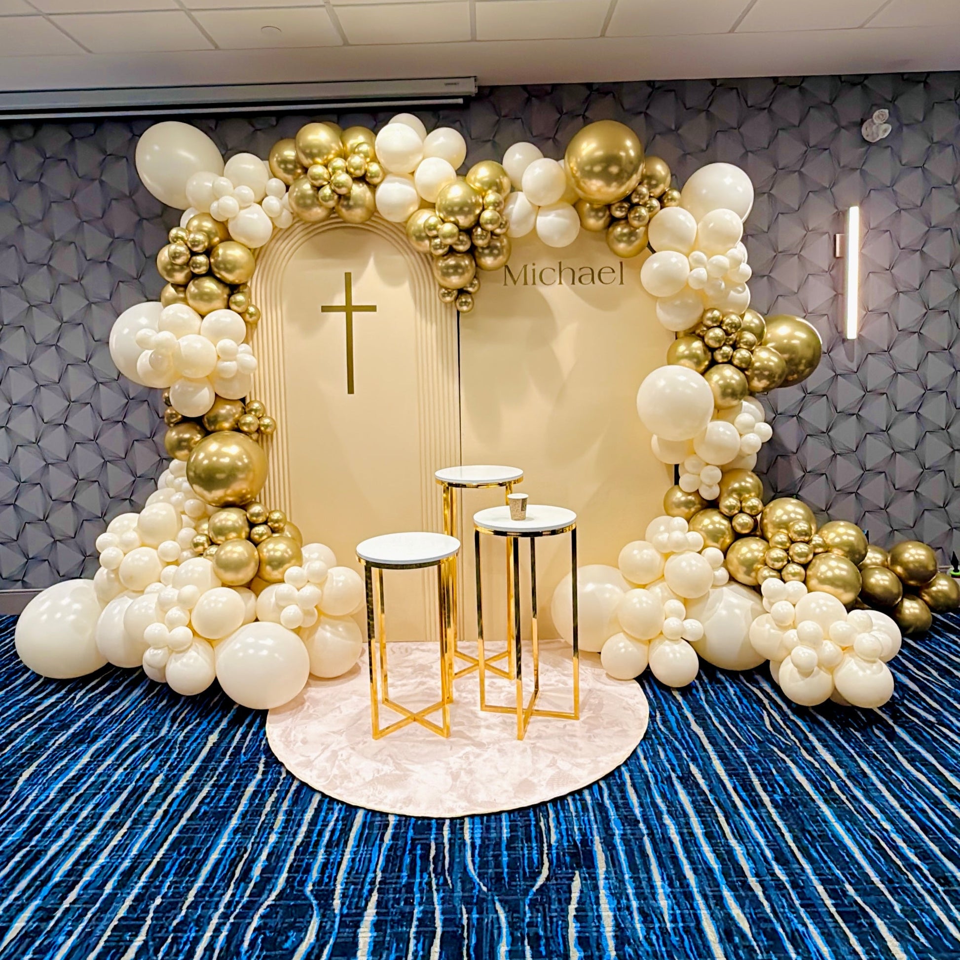 Decorative balloon arch with gold and white balloons in a room with blue carpet and tiled walls.