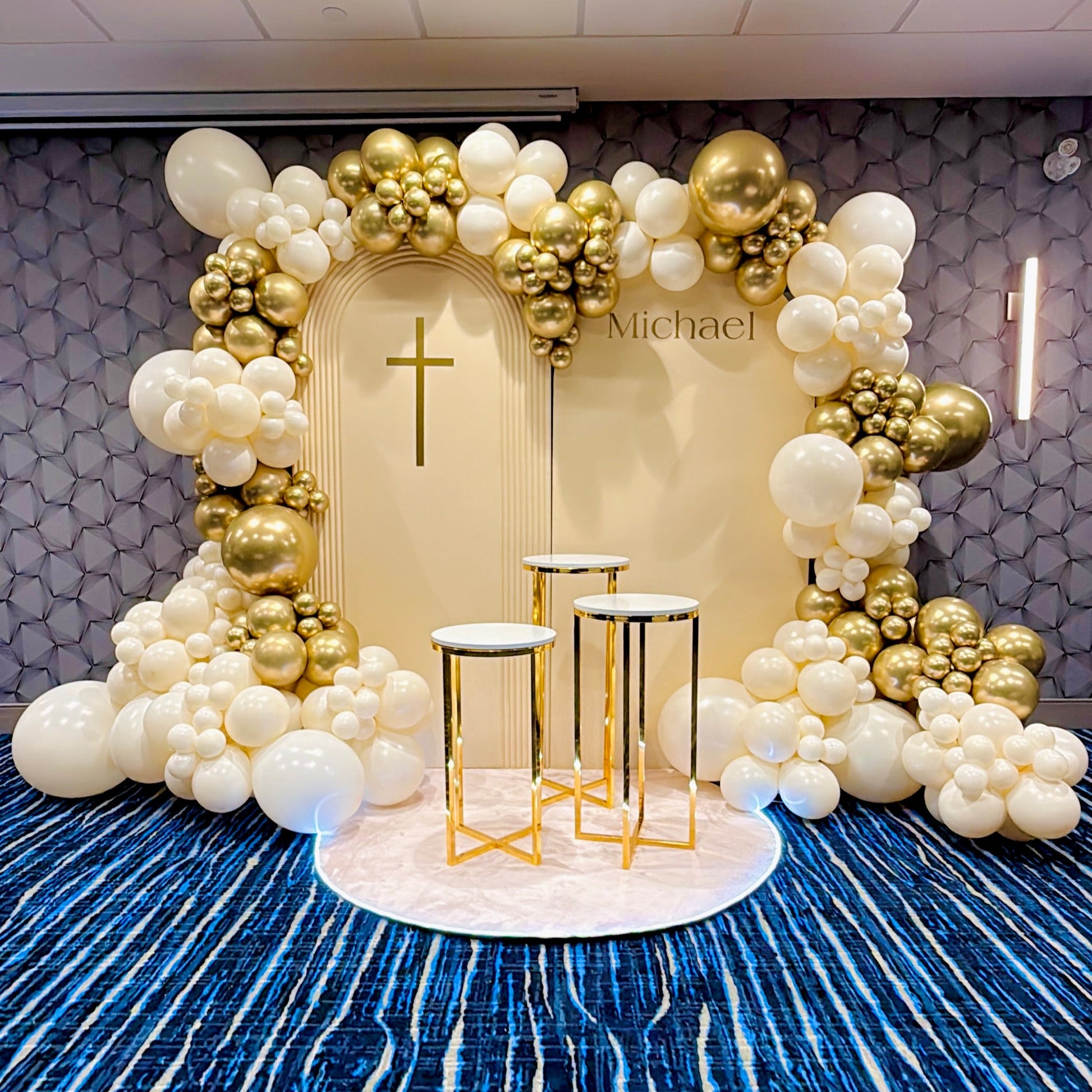 Decorative balloon arch with gold and white balloons in a room with blue patterned carpet.