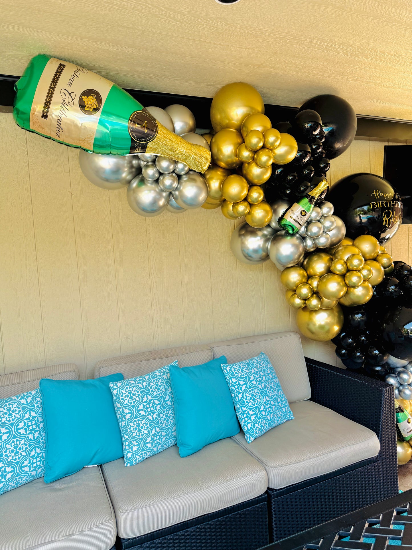 Decorative balloon arch with champagne bottle and balloons above a couch with blue pillows.