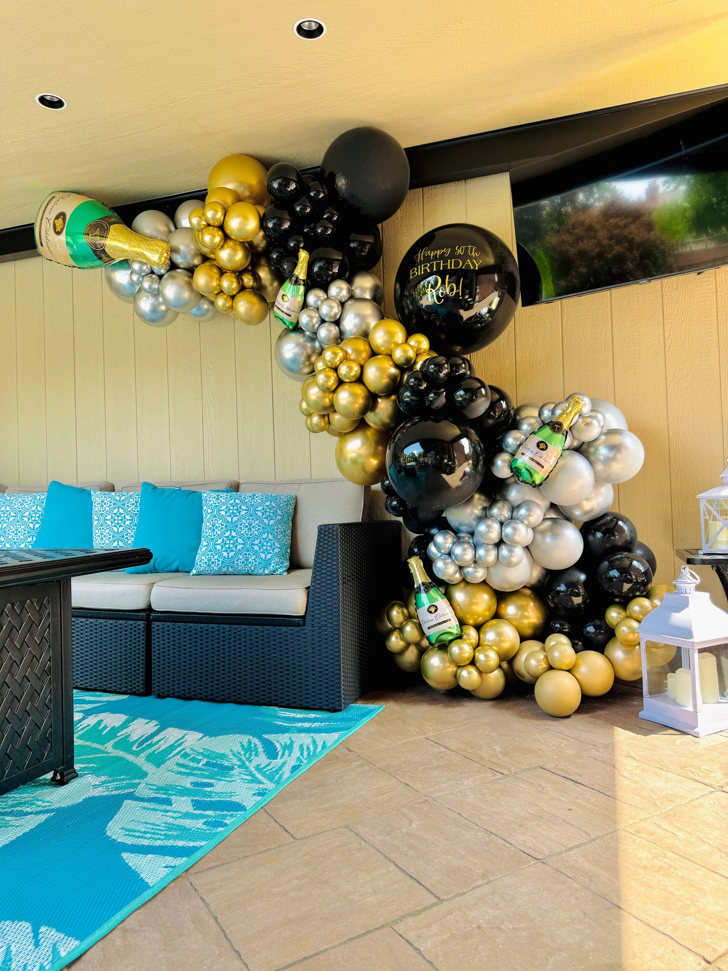 Balloon arch with gold, black, and white balloons in a room with a couch and blue rug.