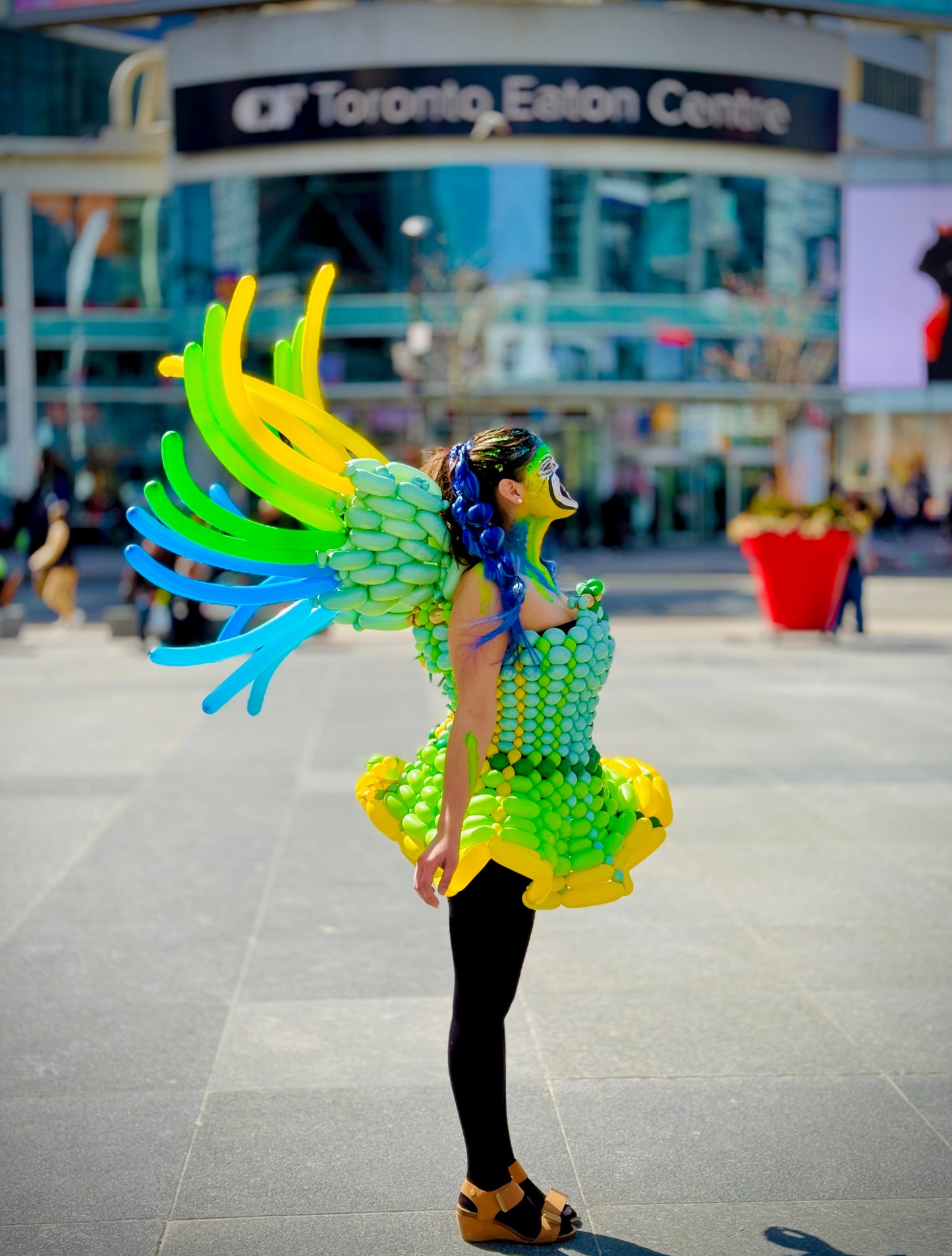 Person in a colorful, green and yellow dress with large wings in front of the Toronto Eaton Centre.