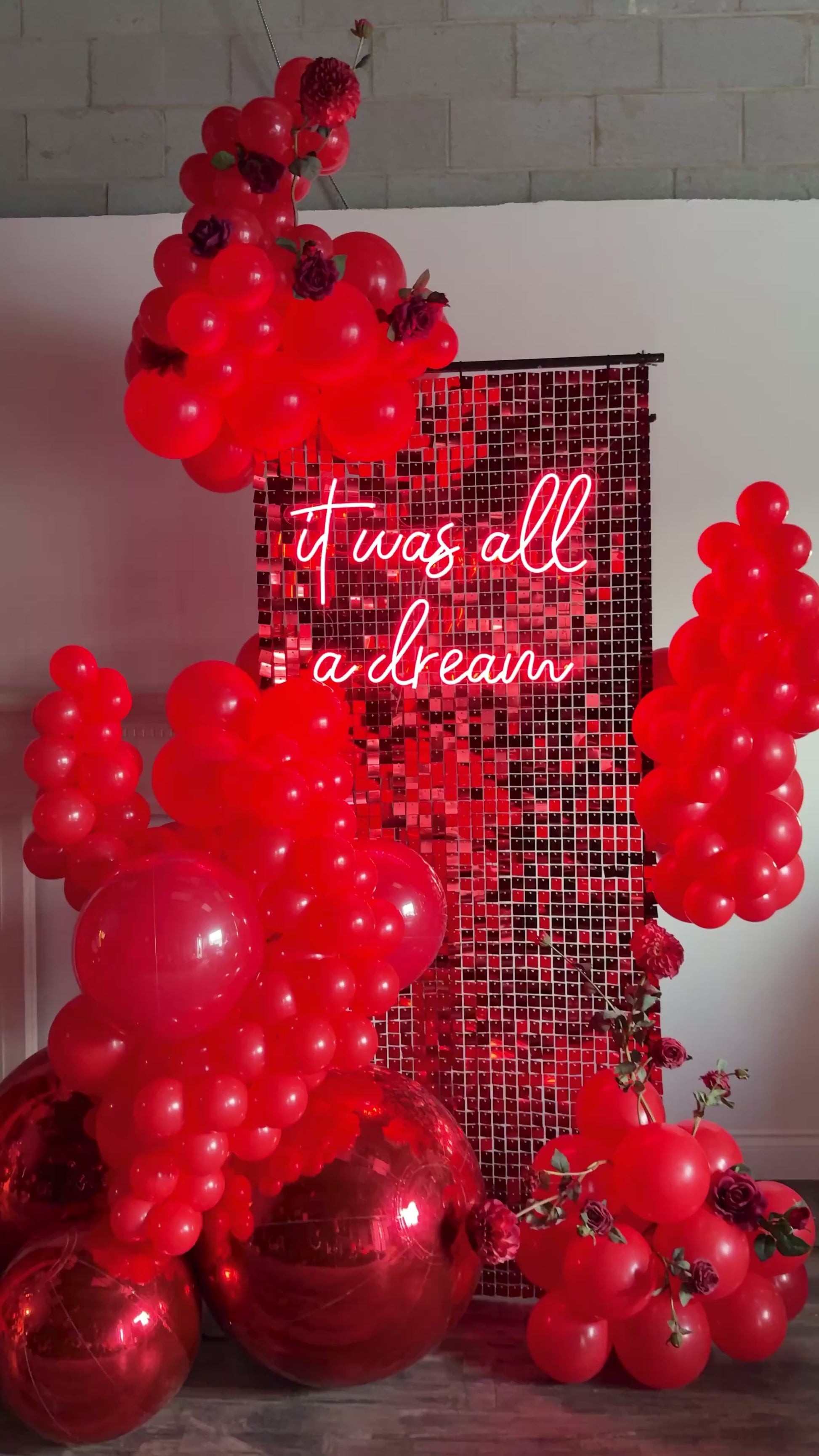 Luxury Red Shimmery Wall Balloon Installation with a red sequin wall, premium balloon garland, LED neon sign, florals, and mirror balls for an elegant event setup.