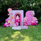 Pink balloon arch and floral decorations on a grassy area with trees in the background