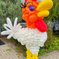 Balloon sculpture of a rooster in an outdoor setting with greenery.