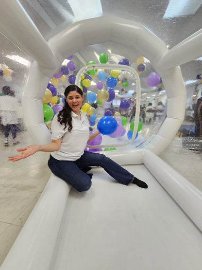 Woman sitting inside a transparent inflatable bubble with colorful balloons in an indoor setting.