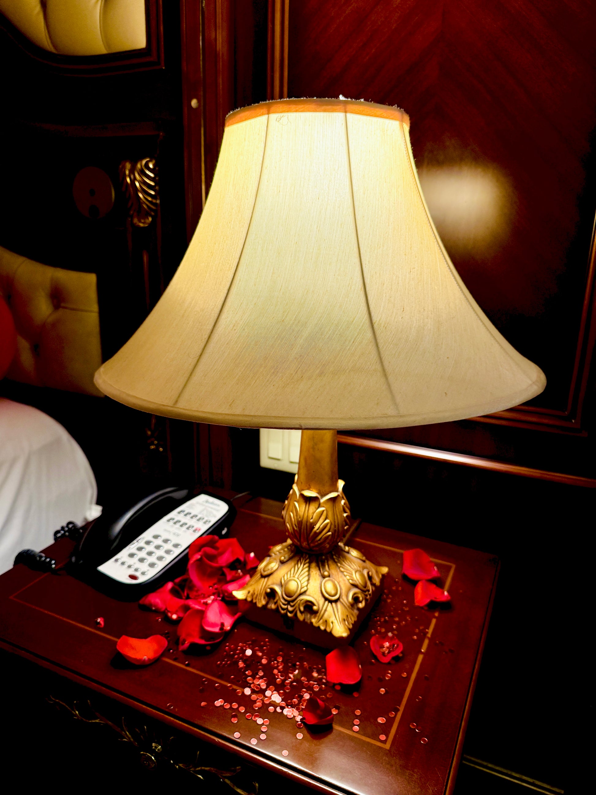 Decorative lamp with a gold base on a wooden table with red flowers and a phone.