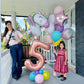 Two women with colorful balloons, including a unicorn and number '5' balloon, on a balcony.