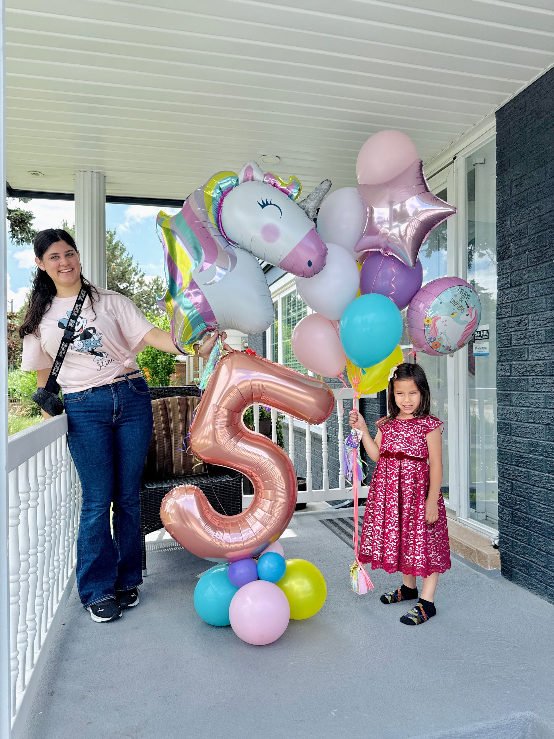 Two women with colorful balloons, including a unicorn and number '5' balloon, on a balcony.