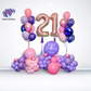 A birthday balloon arrangement featuring large numbered balloons '21' in gold and purple, with a cluster of pink, purple, and white balloons below and a sign with the text 'Happy Birthday Nicky'.