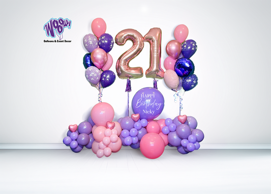 A birthday balloon arrangement featuring large numbered balloons '21' in gold and purple, with a cluster of pink, purple, and white balloons below and a sign with the text 'Happy Birthday Nicky'.