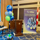 A balloon bouquet arrangement with multiple balloons including a prominent balloon with a logo, displayed in a room with a presentation setup.