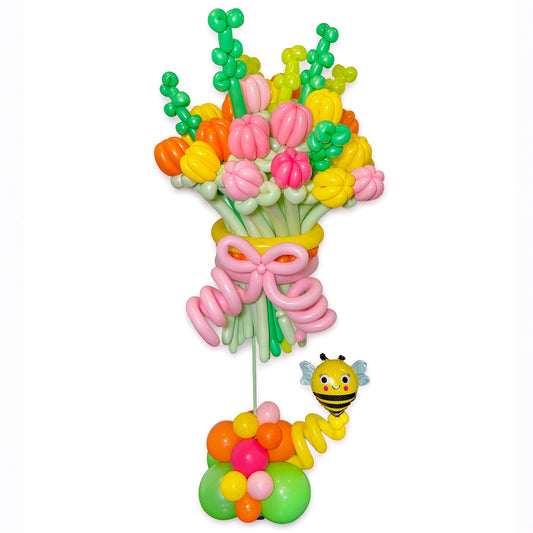 A colorful balloon bouquet decoration with pink, yellow, orange, and green balloons, tied with a pink balloon bow, and a small yellow-and-black bee balloon figure beside it on a plain white background.