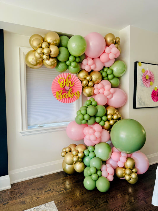 Balloon arch with gold, pink, and green balloons in a room setting.
