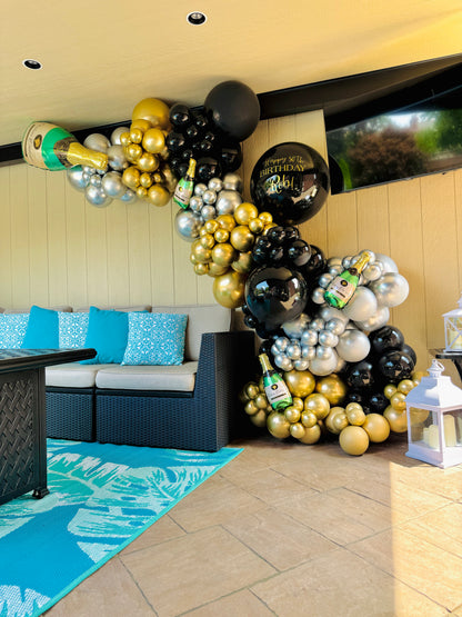 Balloon arch with gold, black, and white balloons in a room with a couch and blue rug.