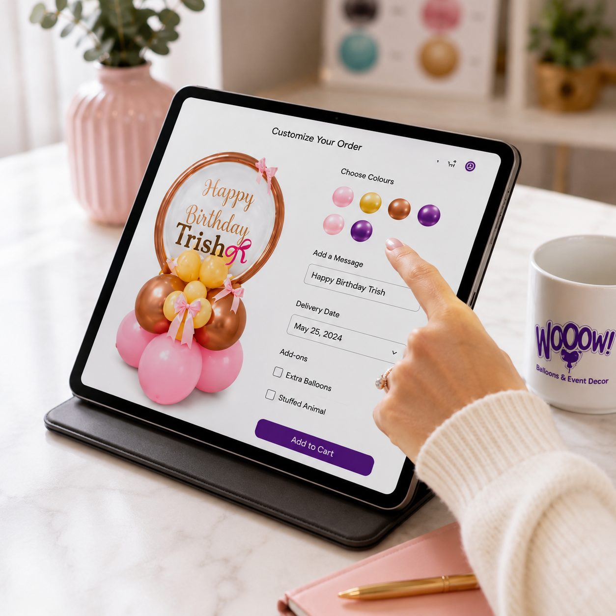 A tablet on a desk shows a birthday balloon order interface with pink, gold, and purple balloons and a custom message. A person's hand is touching the screen.
