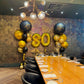 Dining room with a table set for a celebration, featuring gold and black balloons with 'Happy Birthday' and number '80'.