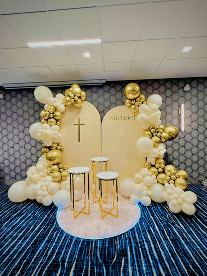 Decorative arch with balloons and a cross on a patterned wall.