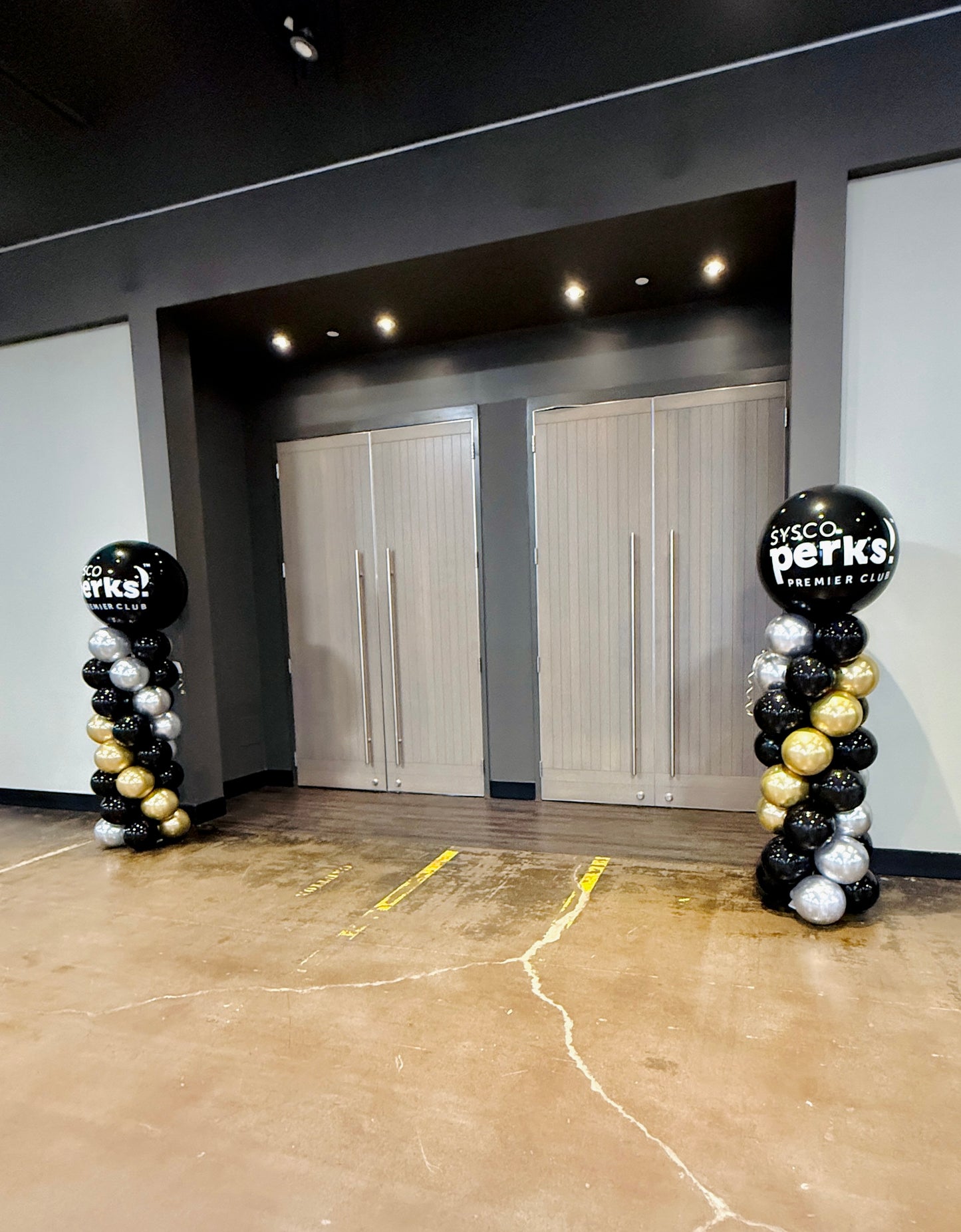 Balloon arches with 'SYSC Perks' branding in a room with white walls and a concrete floor.