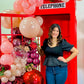 Instagram-worthy red phone booth balloon installation with blush, burgundy, and red balloons, metallic accents, and floral embellishments. A woman strikes a fun pose in front of the setup.