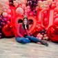 Luxury 'I ❤️ U' balloon installation with a red and blush balloon garland, floral accents, and oversized metallic spheres. A woman poses in front of the glowing setup.