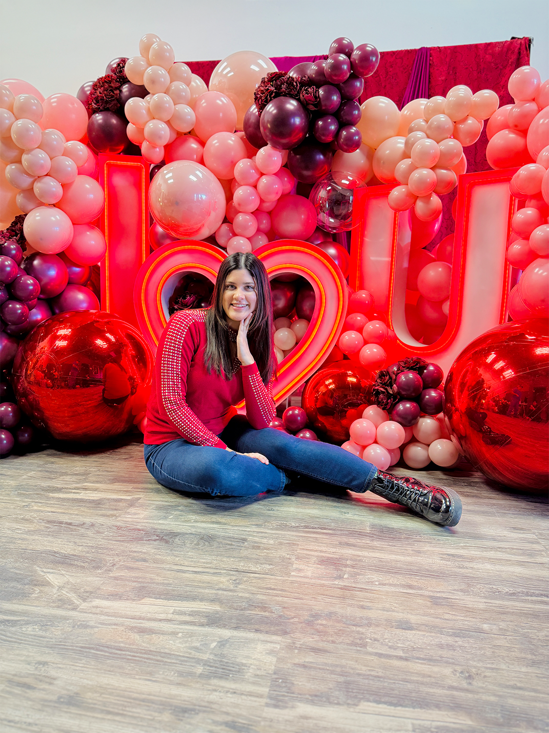 Luxury 'I ❤️ U' balloon installation with a red and blush balloon garland, floral accents, and oversized metallic spheres. A woman poses in front of the glowing setup.