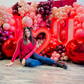 Romantic event decor featuring an illuminated 'I ❤️ U' sign, elegant red and blush balloons, and floral embellishments. A woman sits in front, smiling at the camera.