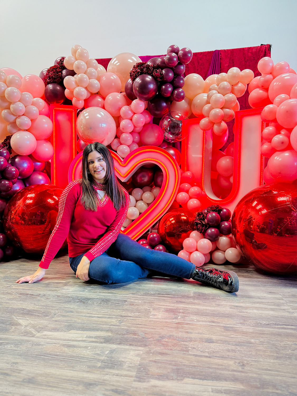 Romantic event decor featuring an illuminated 'I ❤️ U' sign, elegant red and blush balloons, and floral embellishments. A woman sits in front, smiling at the camera.