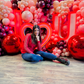 Romantic event decor featuring an illuminated 'I ❤️ U' sign, elegant red and blush balloons, and floral embellishments. A woman sits in front, smiling at the camera.