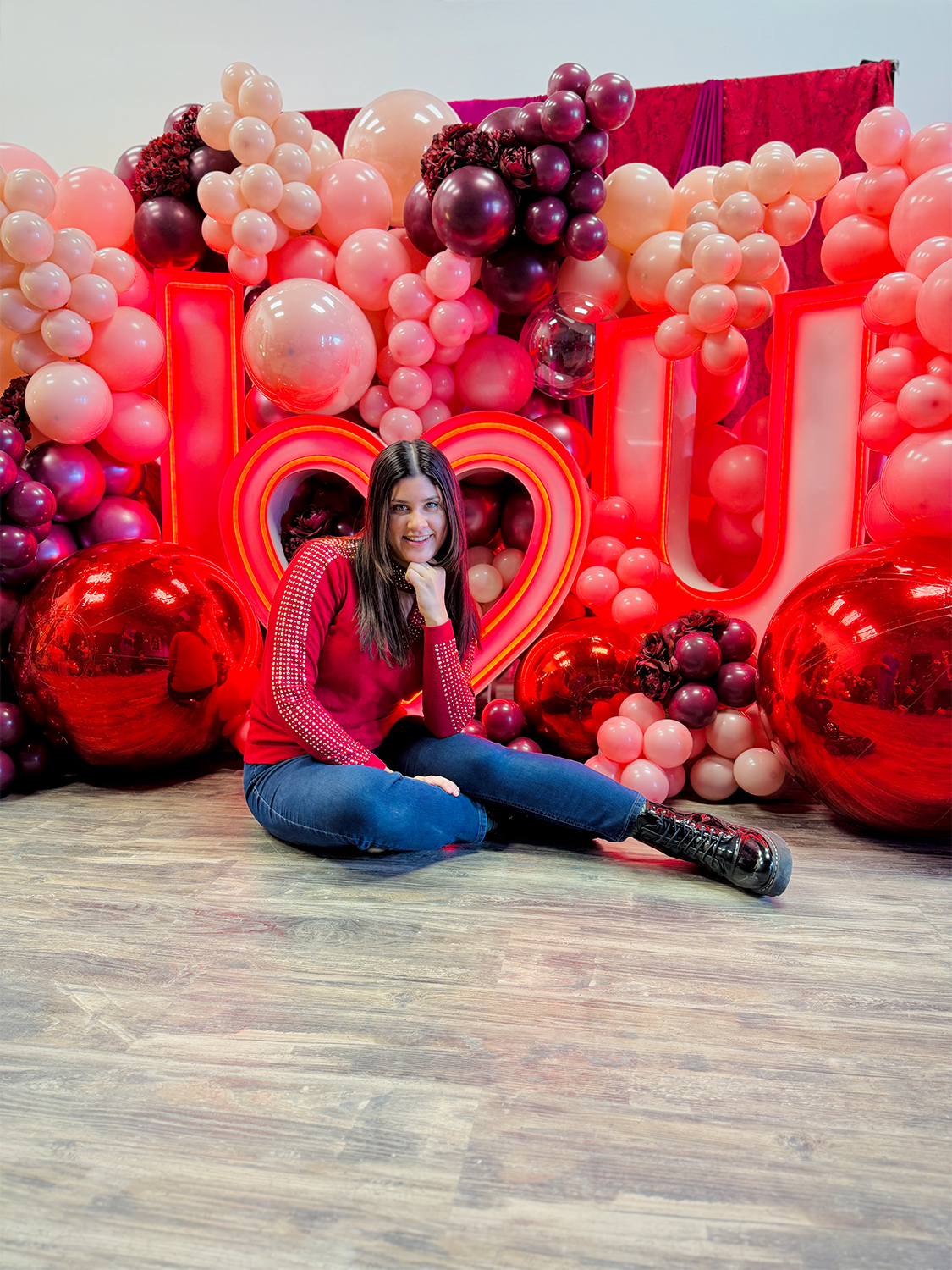 Romantic event decor featuring an illuminated 'I ❤️ U' sign, elegant red and blush balloons, and floral embellishments. A woman sits in front, smiling at the camera.