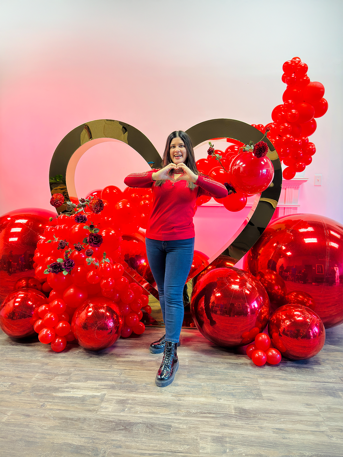 Romantic balloon decor with a gold heart rental, red balloons, floral embellishments, and mirror balls. A woman forms a heart shape with her hands