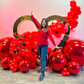 Valentine’s Day balloon setup with a gold heart frame, red balloon clusters, floral decor, and mirror balls. A woman smiles while standing in front of the display