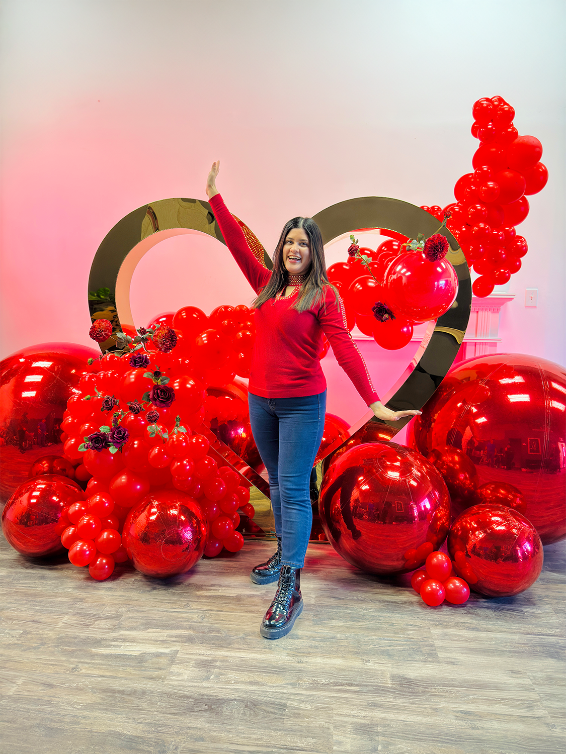 Valentine’s Day balloon setup with a gold heart frame, red balloon clusters, floral decor, and mirror balls. A woman smiles while standing in front of the display