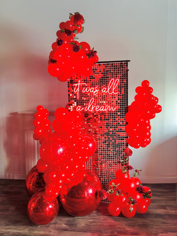 Luxury Red Shimmery Wall Balloon Installation with a red sequin wall, premium balloon garland, LED neon sign, florals, and mirror balls for an elegant event setup.