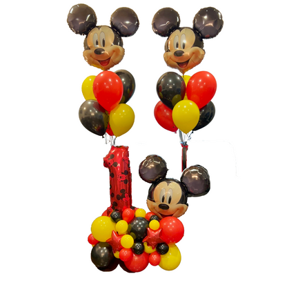 A Mickey Mouse-themed balloon decoration set for a first birthday, featuring two Mickey balloon heads with round black, red, and yellow balloons, a red number 1 balloon, and a base cluster of matching balloons on a white background.
