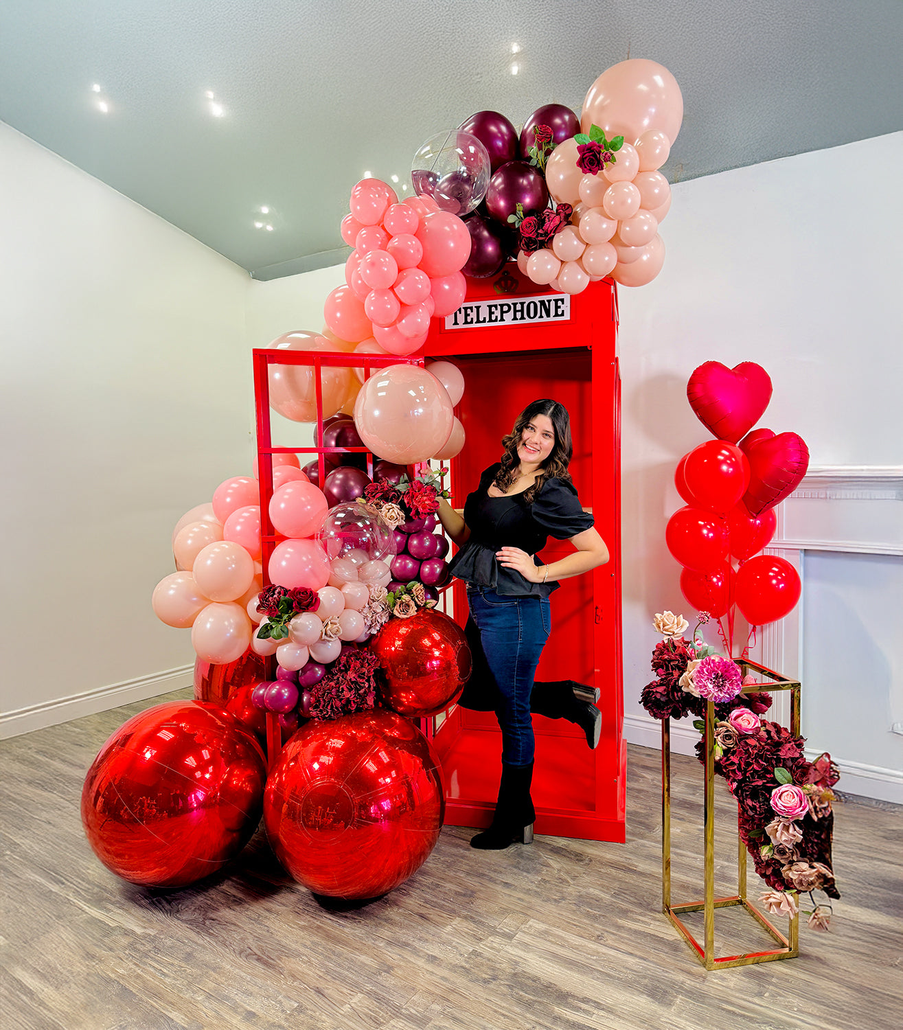 Romantic red phone booth balloon setup featuring blush, burgundy, and red balloons with floral accents. A couple sits inside, creating a cozy and elegant atmosphere.