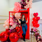 Instagram-worthy red phone booth balloon installation with blush, burgundy, and red balloons, metallic accents, and floral embellishments. A woman strikes a fun pose in front of the setup.