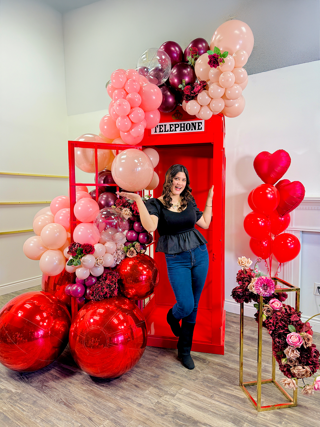 Instagram-worthy red phone booth balloon installation with blush, burgundy, and red balloons, metallic accents, and floral embellishments. A woman strikes a fun pose in front of the setup.