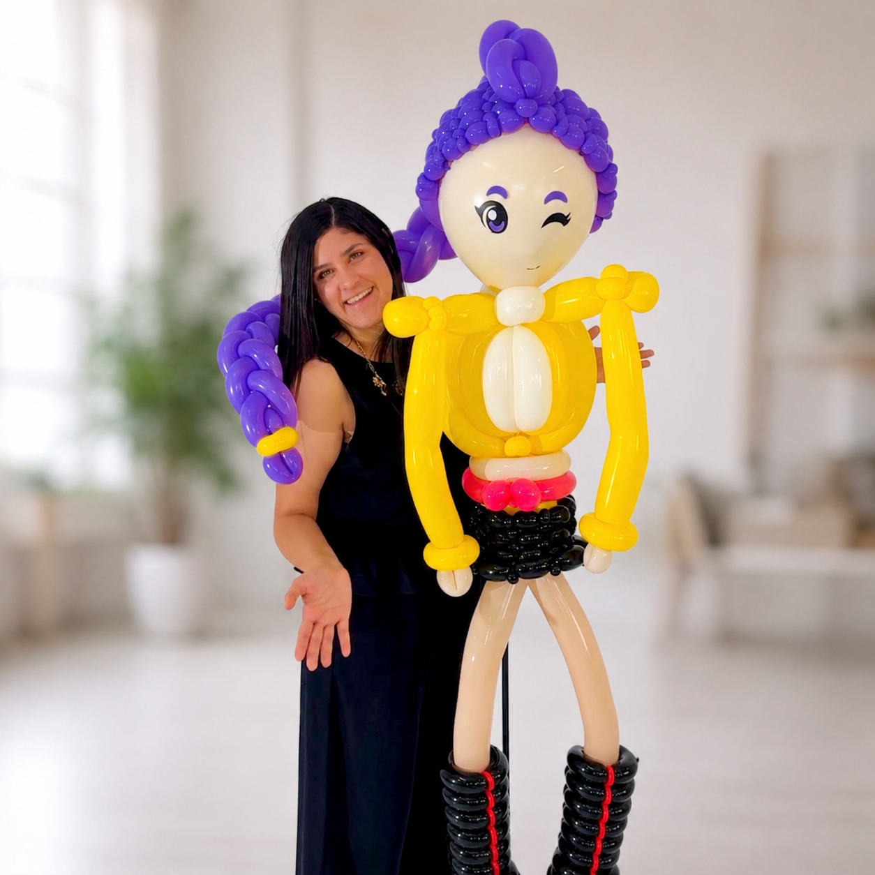 Fransheska Montoya posing beside a handcrafted Rumi Kpop Demon Hunters balloon sculpture with purple braided hair, a yellow outfit, and black boots against a softly blurred indoor background.