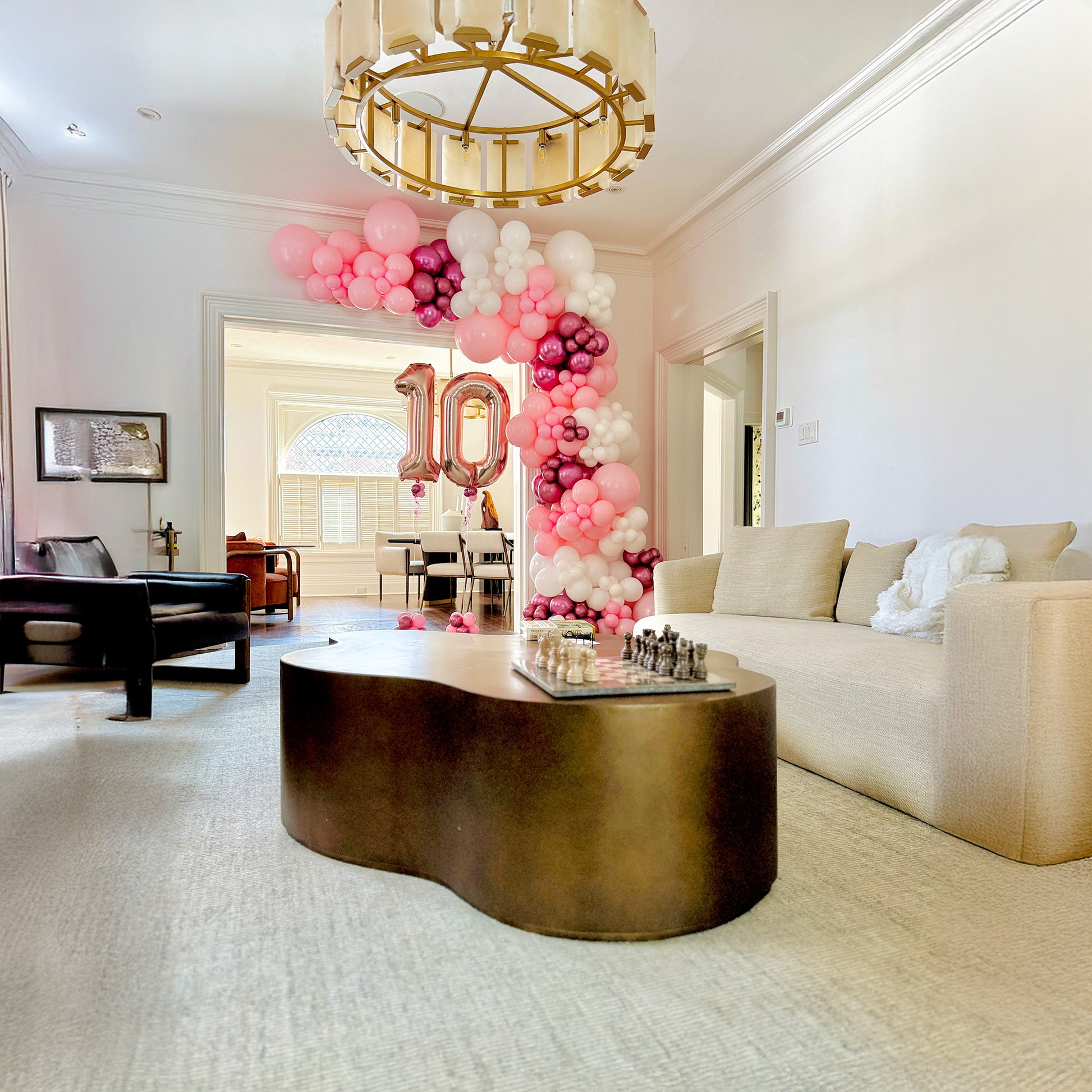 Living room with pink and white balloon arch, '10' balloons, and a wavy coffee table.