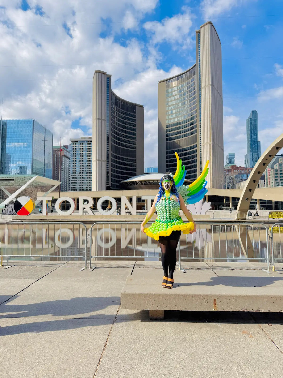 Model wearing a custom green balloon dress with wings beside the Toronto sign at Nathan Phillips Square