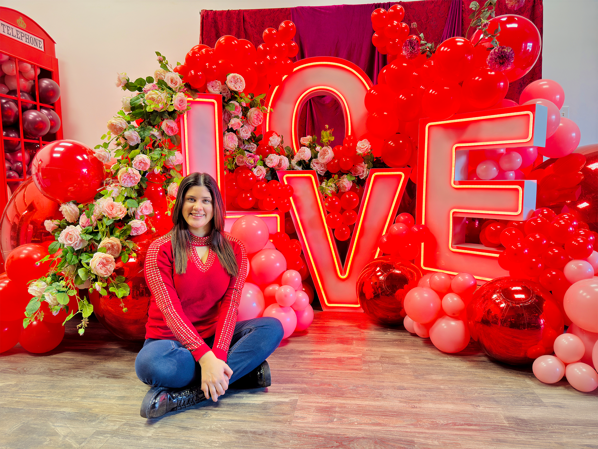 Luxury 'LOVE' marquee letter balloon installation with red and blush balloon garlands, floral accents, and oversized metallic mirror balls. A woman poses in front of the display.