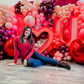 Romantic event decor featuring an illuminated 'I ❤️ U' sign, elegant red and blush balloons, and floral embellishments. A woman sits in front, smiling at the camera.