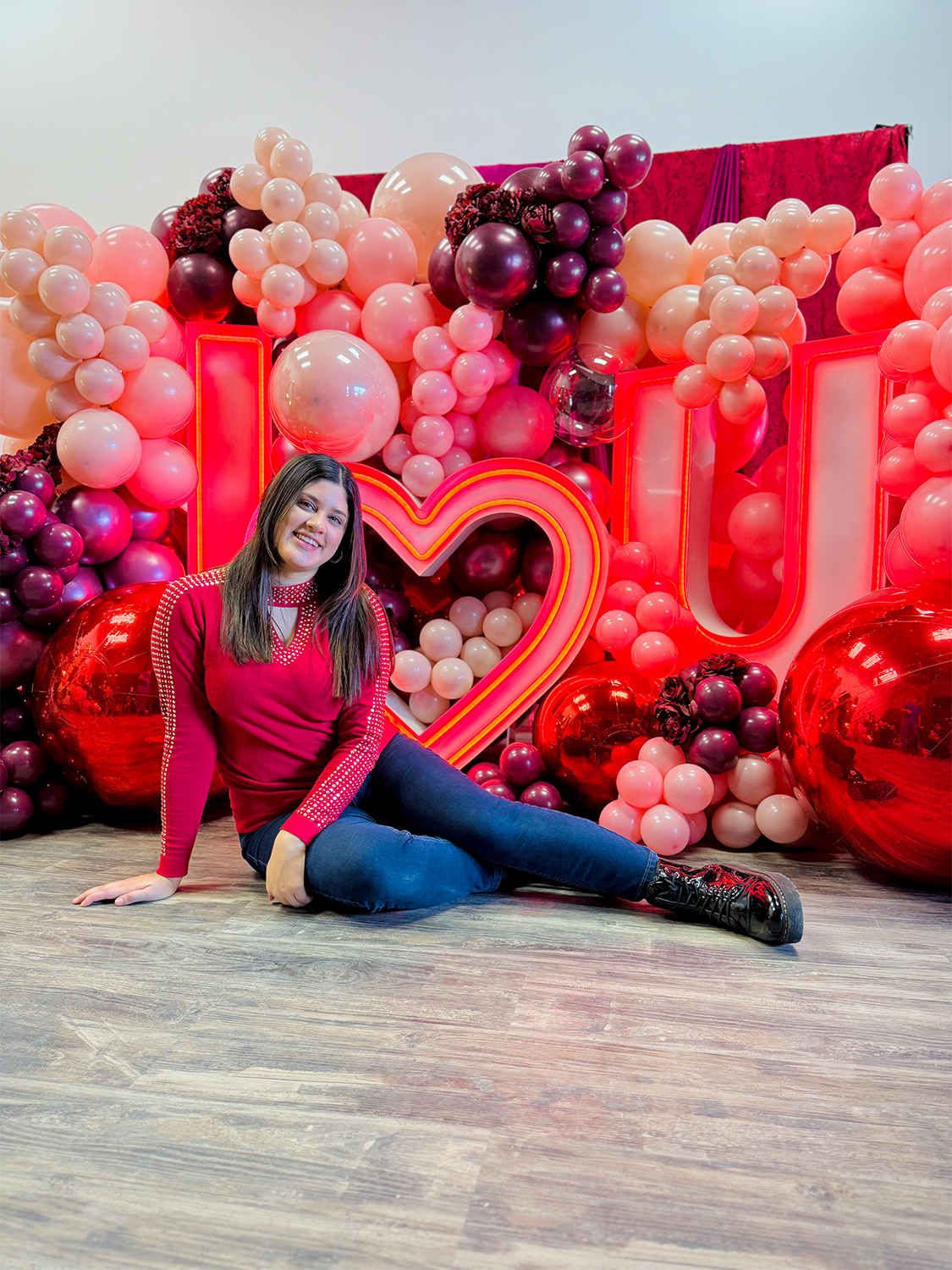 Romantic event decor featuring an illuminated 'I ❤️ U' sign, elegant red and blush balloons, and floral embellishments. A woman sits in front, smiling at the camera.