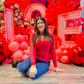 Romantic event decor featuring illuminated 'LOVE' letters, an elegant floral and balloon arrangement, and a red drapery backdrop. A woman sits in front, smiling at the camera.