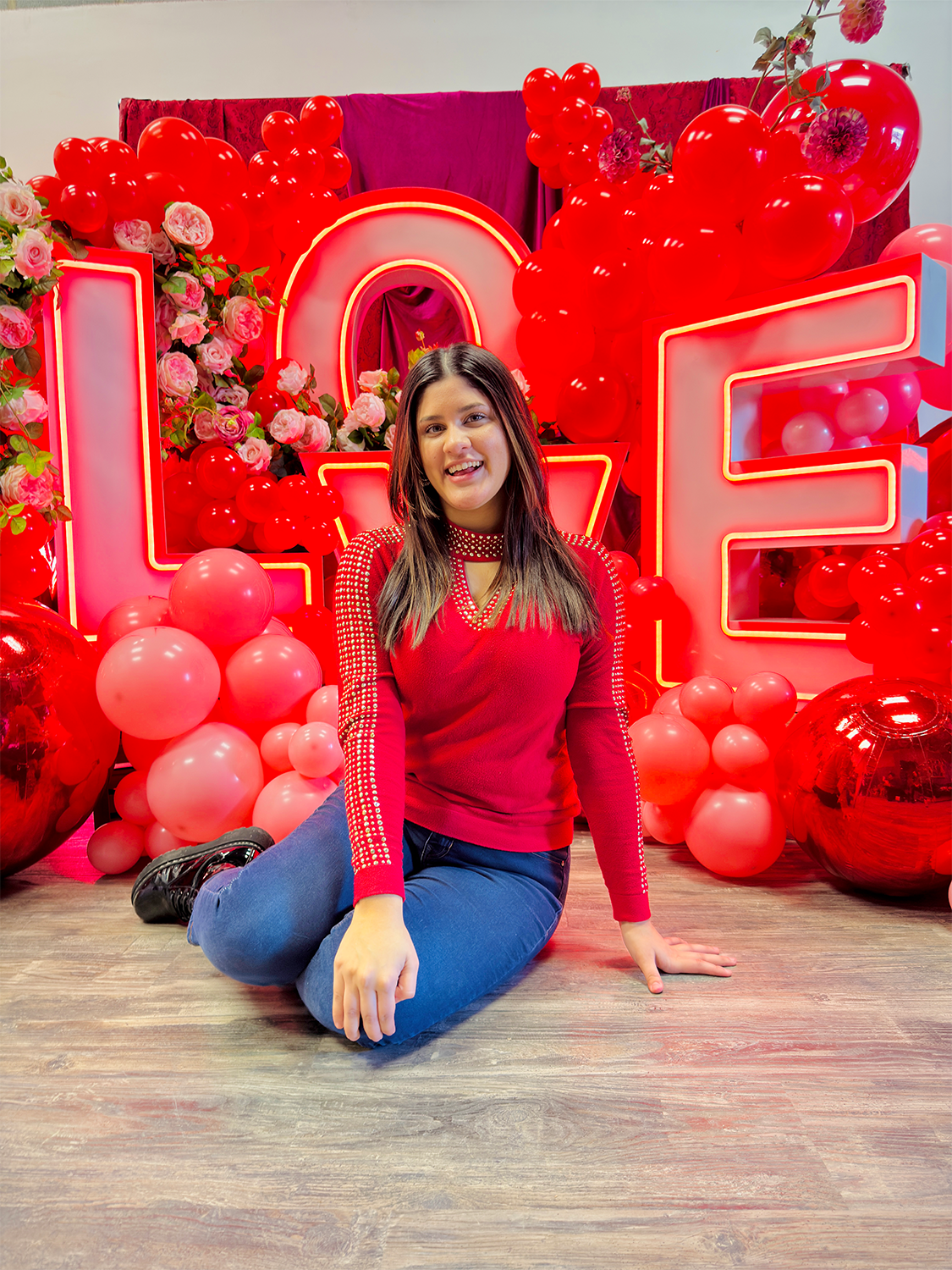 Romantic event decor featuring illuminated 'LOVE' letters, an elegant floral and balloon arrangement, and a red drapery backdrop. A woman sits in front, smiling at the camera.