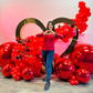 Valentine’s Day balloon setup with a gold heart frame, red balloon clusters, floral decor, and mirror balls. A woman smiles while standing in front of the display