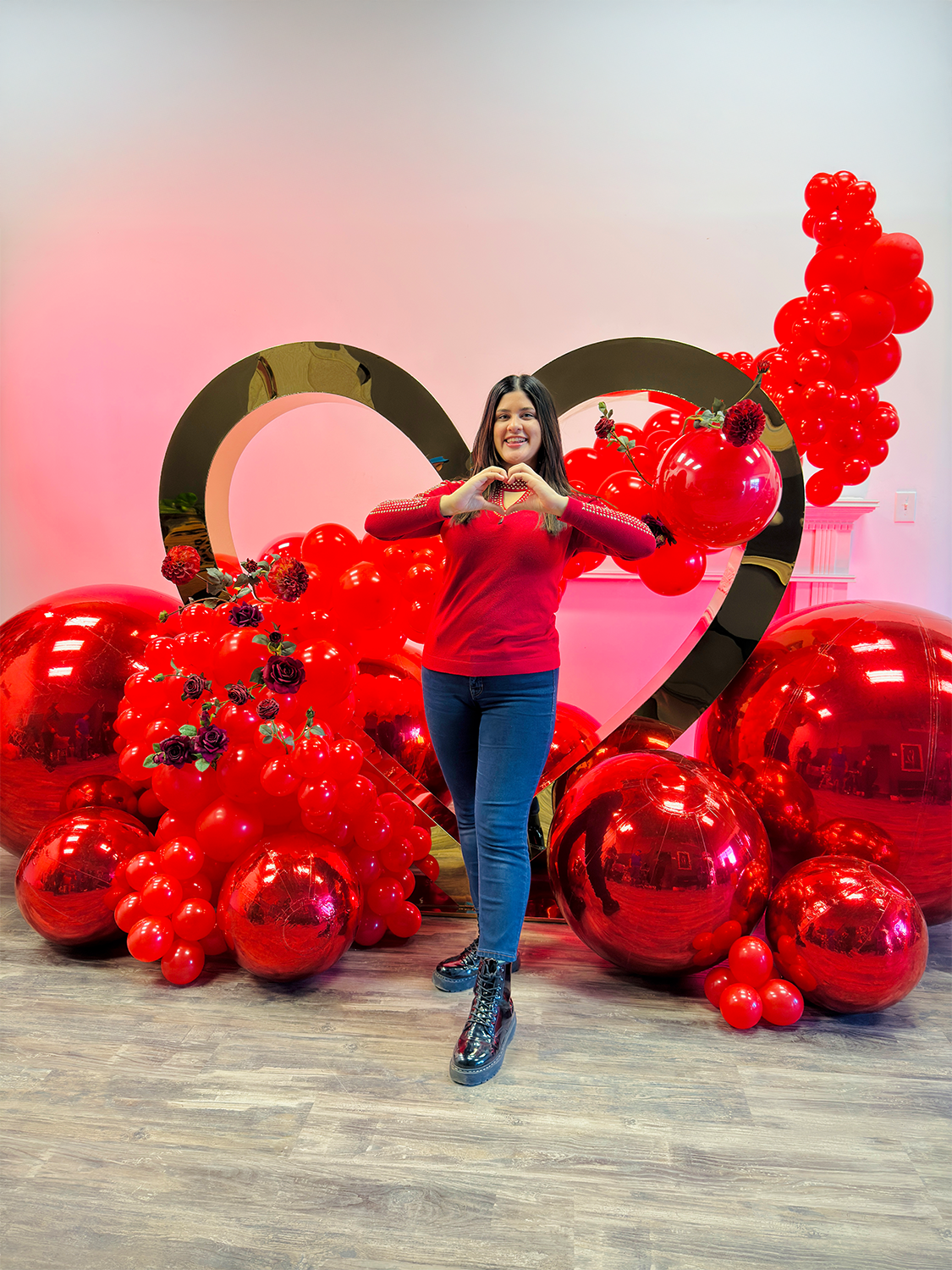 Valentine’s Day balloon setup with a gold heart frame, red balloon clusters, floral decor, and mirror balls. A woman smiles while standing in front of the display